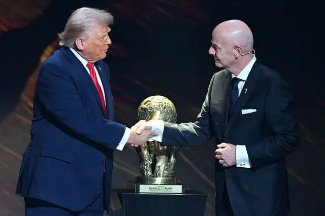 الرئيس الأمريكي دونالد ترامب يصافح رئيس الاتحاد الدولي لكرة القدم (ألفيفا) جياني إنفانتينو أثناء تسلمه "جائزة الفيفا للسلام"، وذلك خلال مراسم سحب قرعة بطولة كأس العالم لكرة القدم 2026، ويرتدي ترامب سترة زرقاء وقميصاً أبيض وربطة عنق حمراء، بينما يرتدي إنفانتينو سترة سوداء وقميصاً أبيض وربطة عنق زرقاء.