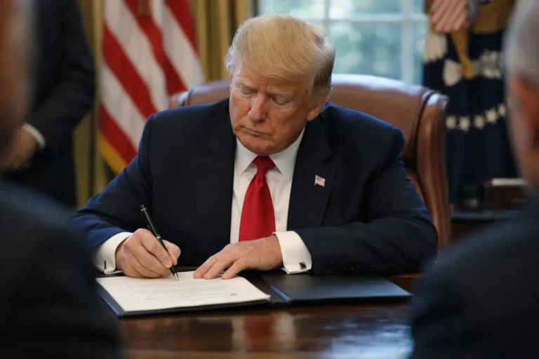 Donald Trump signs a document at the Oval Office desk.